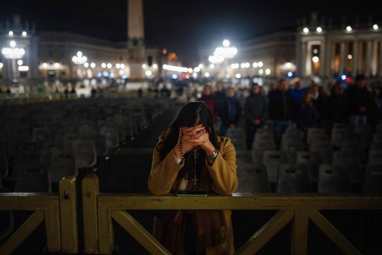 A imagem mostra uma mulher em um momento de reflexão ou tristeza, com as mãos cobrindo o rosto. Ela está em frente a uma grade, em uma praça iluminada à noite, onde há várias cadeiras vazias. Ao fundo, é possível ver um obelisco e outras pessoas em pé, também em silêncio. A iluminação da praça cria um ambiente solene.