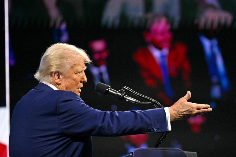 A imagem mostra um homem de cabelo loiro e vestido com um terno azul, falando em um microfone. Ele está gesticulando com a mão direita, enquanto ao fundo há uma tela com a imagem de pessoas assistindo ao evento. A iluminação é focada no orador, destacando sua figura em contraste com o fundo mais escuro.