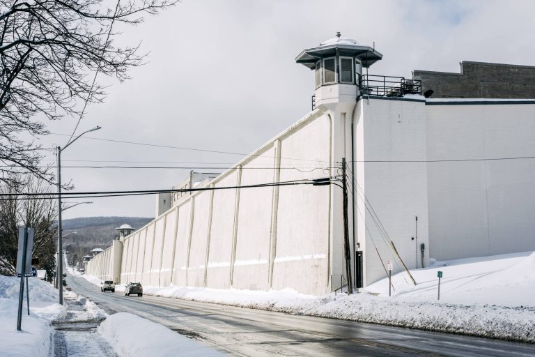 A imagem mostra uma prisão com uma alta parede branca e uma torre de vigilância no topo. A cena é em um dia nublado, com neve acumulada ao longo da estrada e nas laterais. Há um carro passando na estrada em frente à prisão, que é cercada por árvores sem folhas.