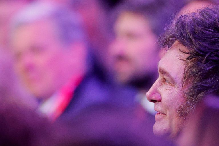 A imagem mostra o perfil de um homem sorrindo, com cabelo escuro e ondulado. Ao fundo, há outras pessoas desfocadas, com um ambiente iluminado em tons de roxo e azul. O homem parece estar em um evento social ou formal.