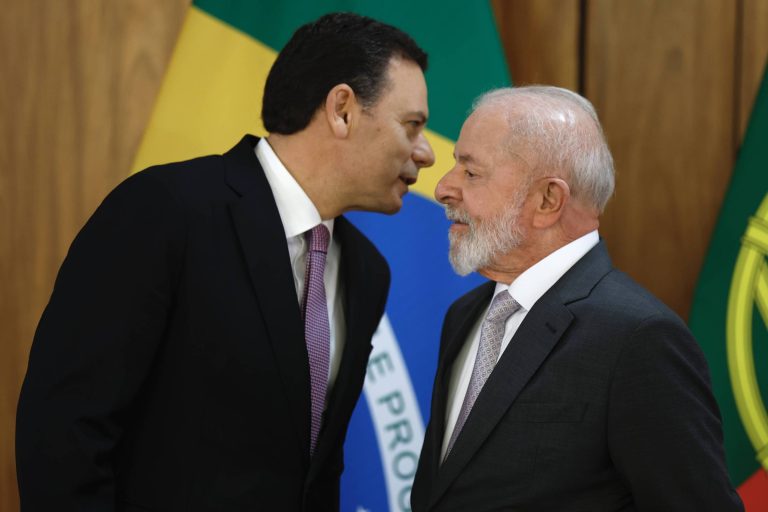 Dois homens estão em um momento de conversa próxima, com expressões sérias. Ao fundo, há bandeiras do Brasil, uma com as cores verde e amarelo e outra com o brasão nacional. O homem à esquerda usa um terno escuro com uma gravata clara, enquanto o homem à direita está vestido com um terno escuro e uma gravata prateada.