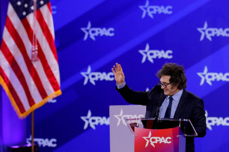 A imagem mostra um homem em um palco durante o evento CPAC, com uma bandeira dos Estados Unidos ao fundo. O homem está de pé, com um microfone à sua frente, acenando com a mão direita. O fundo é decorado com um padrão azul e o logotipo do CPAC em branco.