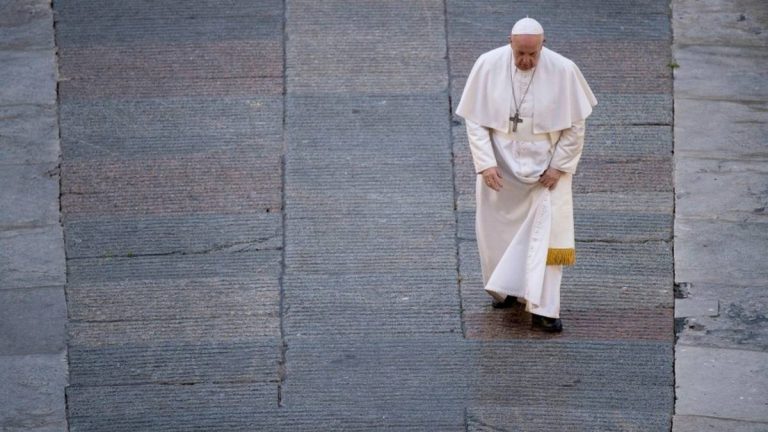 A imagem mostra um homem vestido com uma vestimenta branca, que parece ser um papa, caminhando em um espaço pavimentado. Ele está ajustando a parte inferior de sua vestimenta enquanto se move. O chão é composto por pedras em tons de cinza e há uma iluminação suave.
