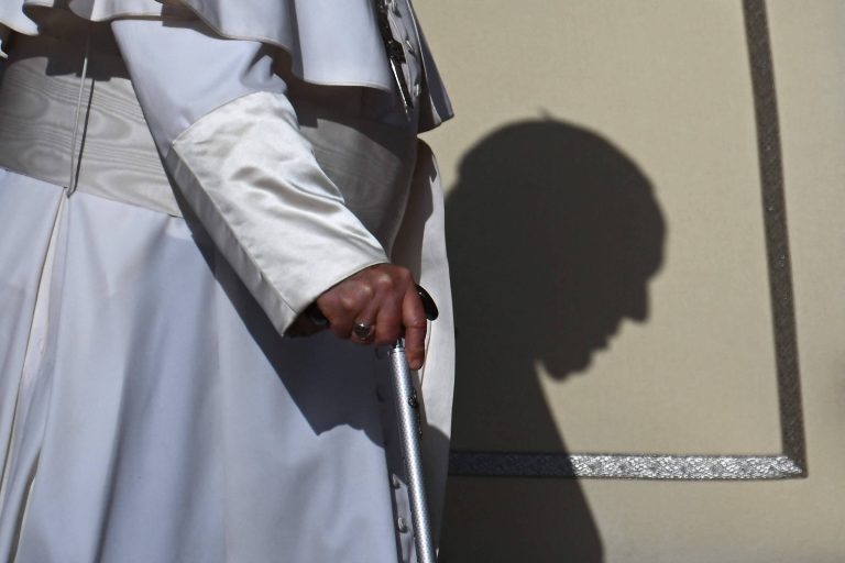 A imagem mostra uma mão segurando uma bengala, com uma figura vestida de branco ao fundo. A sombra da figura é projetada na parede, criando um contorno que sugere um rosto. A iluminação destaca a bengala e a vestimenta branca, enquanto a parede é de uma cor clara.