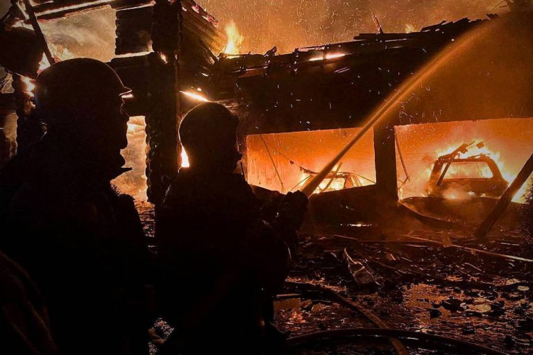 A imagem mostra bombeiros lutando contra um incêndio em uma estrutura em chamas. As chamas são intensas, iluminando a cena com um brilho laranja. Os bombeiros estão usando mangueiras para combater o fogo, enquanto a estrutura queimada está parcialmente colapsada. É possível ver veículos danificados nas proximidades.