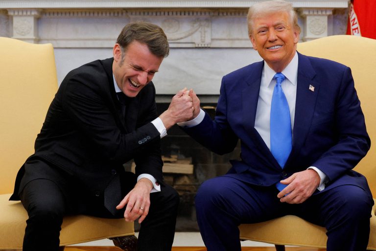 Na imagem, duas pessoas estão sentadas em cadeiras em um ambiente formal. À esquerda, um homem de terno escuro e gravata clara está sorrindo e rindo, enquanto à direita, um homem de terno azul claro e gravata azul escura também está sorrindo. Eles estão se cumprimentando com um aperto de mão, em um gesto de camaradagem. Ao fundo, há uma lareira e uma decoração clássica.