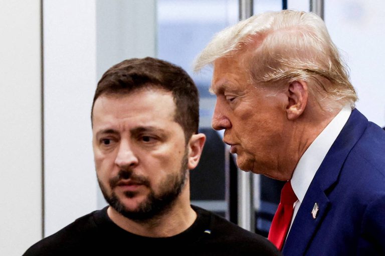 A imagem mostra dois homens em um ambiente interno. Um deles está de perfil, com cabelo escuro e barba, vestindo uma camiseta preta. O outro homem, com cabelo loiro e penteado, está inclinado em direção ao primeiro, parecendo falar com ele. O fundo é desfocado, mas sugere um ambiente de reunião.