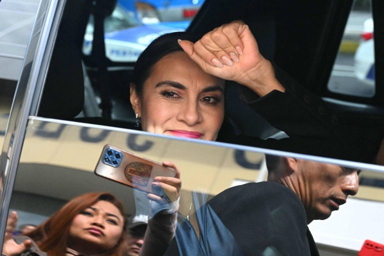 A imagem mostra uma mulher acenando com a mão de dentro de um carro. Ela tem cabelo escuro e usa roupas escuras. Ao fundo, é possível ver outras pessoas e um veículo da polícia. A mulher parece estar sorrindo e há um reflexo de uma pessoa segurando um celular na janela do carro.