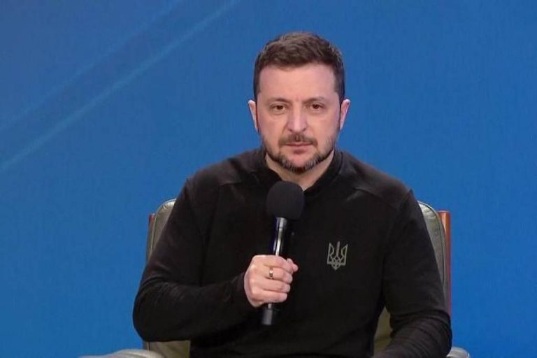 Um homem sentado em uma cadeira, segurando um microfone. Ele está usando uma camiseta preta com um símbolo em destaque. O fundo é azul, e ele parece estar em um ambiente de entrevista ou conferência.
