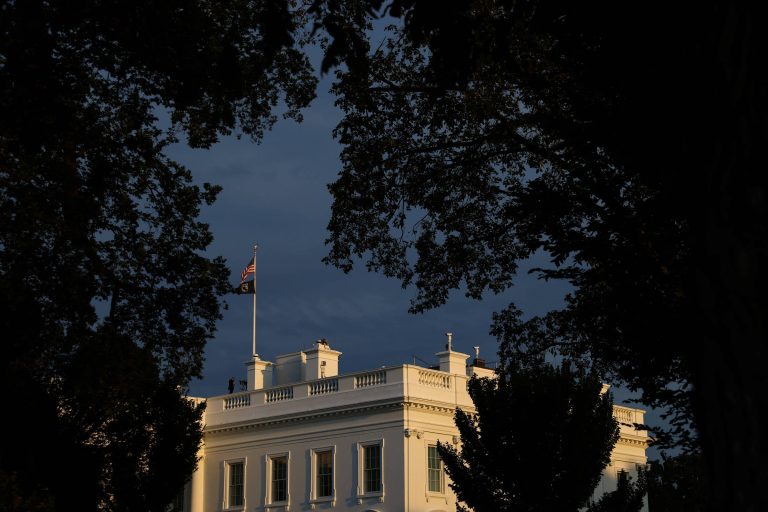 A imagem mostra a Casa Branca parcialmente visível entre as árvores. O céu está escuro, sugerindo que é ao entardecer. Uma bandeira está hasteada no topo do edifício.