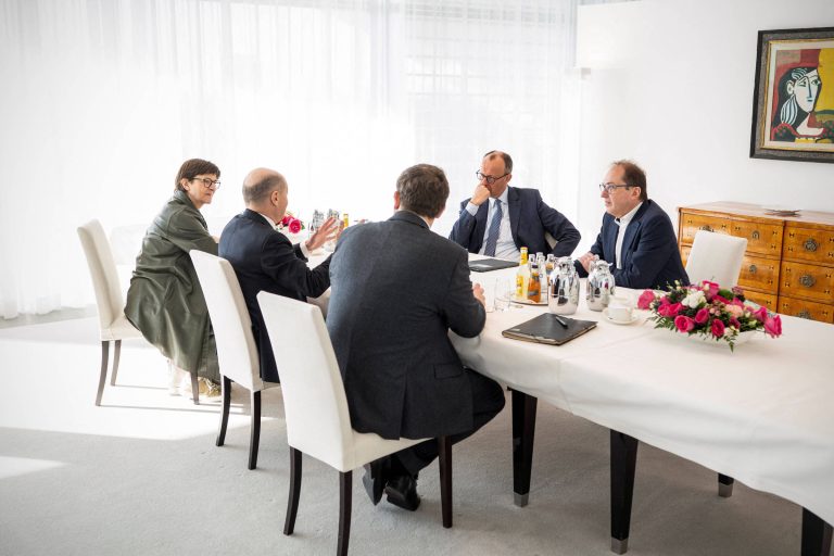 A imagem mostra uma reunião em uma sala iluminada por luz natural. Seis pessoas estão sentadas ao redor de uma mesa retangular coberta com uma toalha branca. Há flores em um vaso no centro da mesa e garrafas de bebida. As pessoas estão vestidas formalmente, algumas estão conversando e outras ouvindo. Ao fundo, há uma obra de arte na parede e um móvel de madeira.