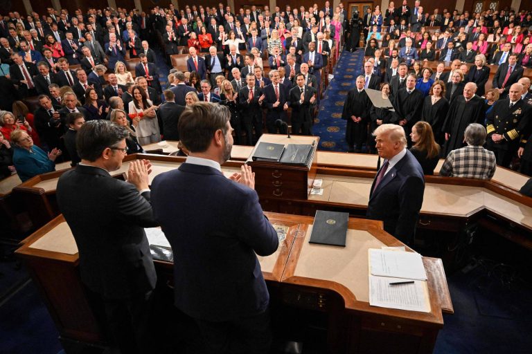 A imagem mostra uma sessão do Congresso dos Estados Unidos, com uma grande audiência. No centro, um homem de terno escuro está se dirigindo ao público, enquanto várias pessoas aplaudem. Há muitos membros do Congresso e outras autoridades presentes, todos em pé, em um ambiente formal. O cenário é bem iluminado, com uma disposição de cadeiras em fileiras e uma mesa na frente do orador.