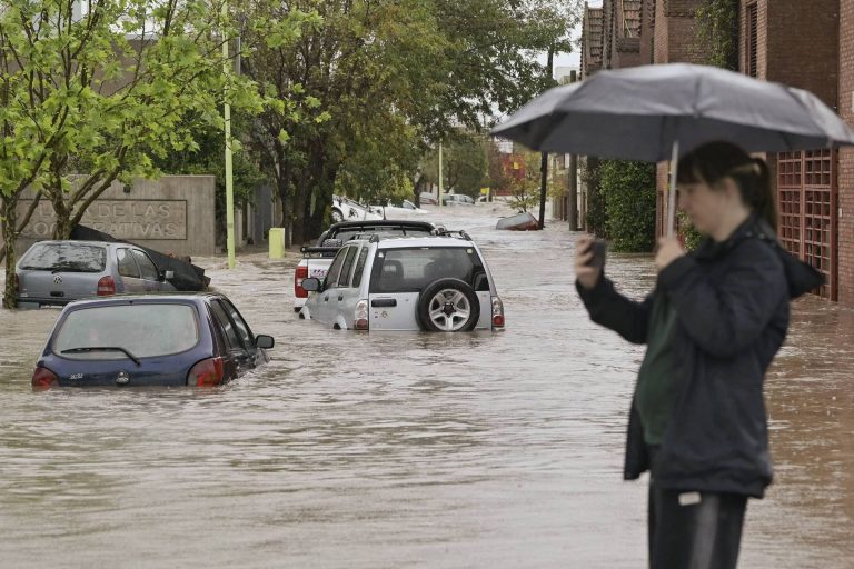 A imagem mostra uma rua inundada, com vários carros submersos em água. Uma mulher está em primeiro plano, segurando um guarda-chuva e usando um celular. Ao fundo, há árvores e prédios, e a água parece cobrir a maior parte da via.