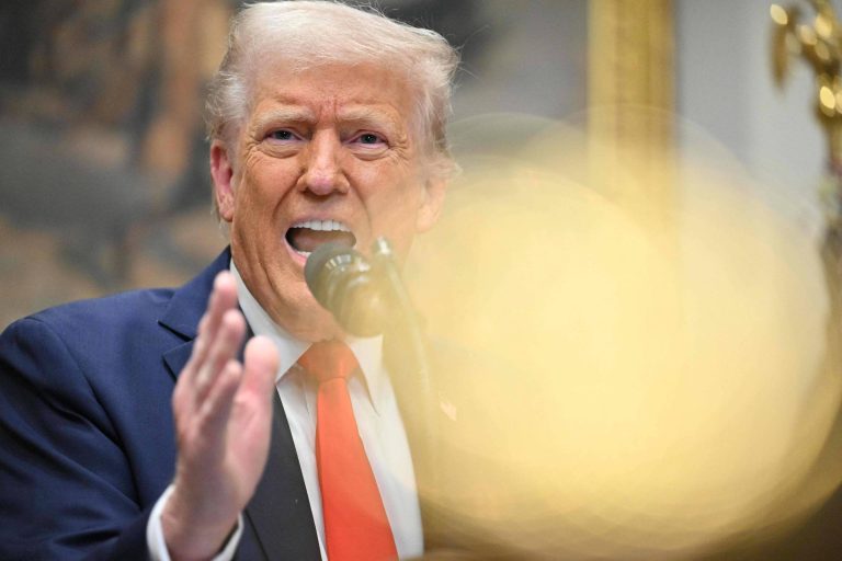 Um homem com cabelo loiro e pele clara está falando em um microfone. Ele usa um terno escuro com uma gravata vermelha. A imagem está levemente desfocada na parte frontal, com um brilho amarelo. Ao fundo, há uma parede decorada e outros elementos que não são claramente visíveis.