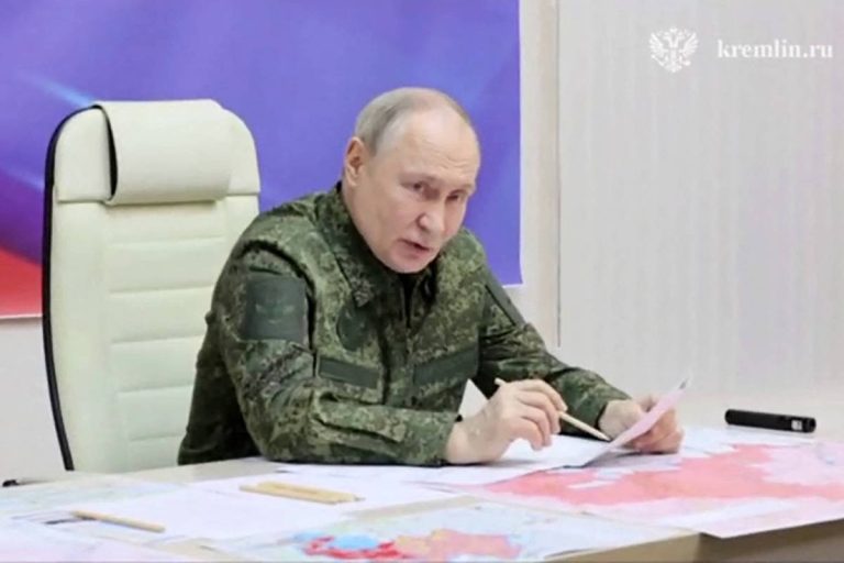 Um homem sentado em uma cadeira branca, vestindo um uniforme militar camuflado, está analisando documentos sobre uma mesa coberta com mapas. Ao fundo, há um painel com cores roxas e azuis. O ambiente parece ser uma sala de reuniões.