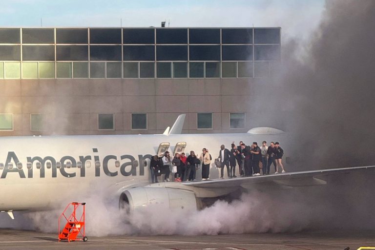 A imagem mostra um avião da American Airlines com fumaça saindo de um dos motores. Um grupo de pessoas está em pé na asa do avião, enquanto a fumaça se espalha ao redor. Ao fundo, há um edifício com janelas.