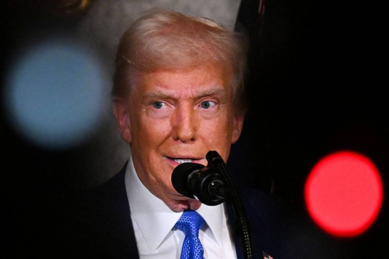 A imagem mostra um homem com cabelo loiro e uma gravata azul, falando em um microfone. O fundo é desfocado, com luzes vermelhas e brancas visíveis. O homem parece estar em um evento formal.