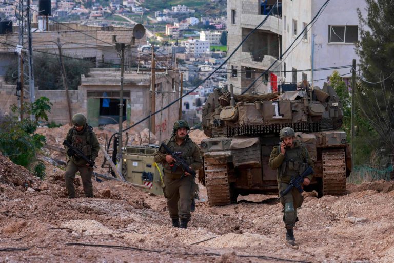 A imagem mostra soldados caminhando em uma área urbana, com um tanque de guerra ao fundo. Os soldados estão vestidos com uniformes militares e portando armas. O cenário é montanhoso, com edifícios e casas visíveis ao fundo, além de fios elétricos e vegetação ao lado da estrada de terra.