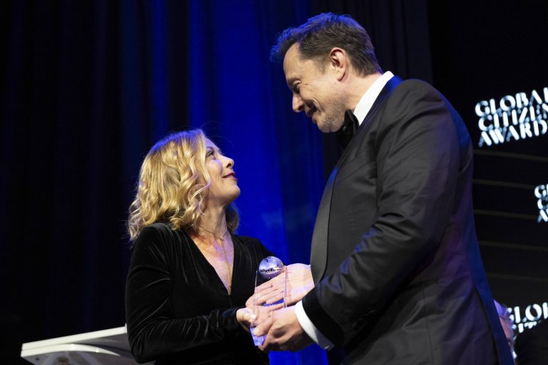 A imagem mostra duas pessoas em uma cerimônia de premiação. Uma mulher com cabelo loiro e ondulado, vestindo um vestido preto, está olhando para um homem que usa um terno preto. O homem está segurando um prêmio em forma de cristal, enquanto a mulher parece estar recebendo o prêmio. O fundo é escuro com luzes azuis e há um telão exibindo o texto