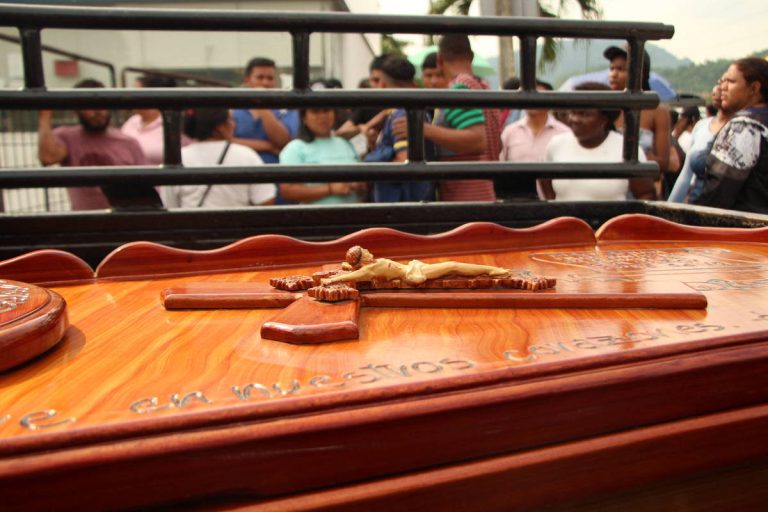 A imagem mostra uma cruz de madeira com uma figura de Cristo crucificado em primeiro plano. Ao fundo, há uma multidão de pessoas, algumas visíveis, observando. A cena parece ser de um evento religioso ou uma cerimônia, com um ambiente ao ar livre.