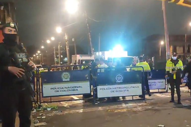 A imagem mostra uma cena noturna em Bogotá, Colômbia, com vários policiais em uniformes, alguns armados, em frente a barreiras de contenção. As barreiras têm placas que indicam
