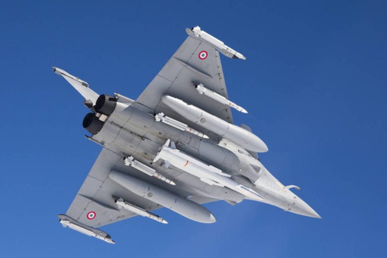 Foto mostra avião militar por baixo, voando num céu azul. Ele está armado com um grande míssil no ventre, além de seis mísseis menores na asa e fuselagem