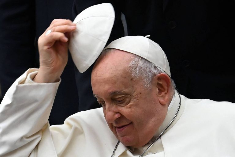 Imagem de um homem idoso, vestido com vestes brancas, que está levantando uma mitra branca da cabeça. Ele tem um sorriso suave e está em um ambiente com pessoas ao fundo, vestidas de preto.