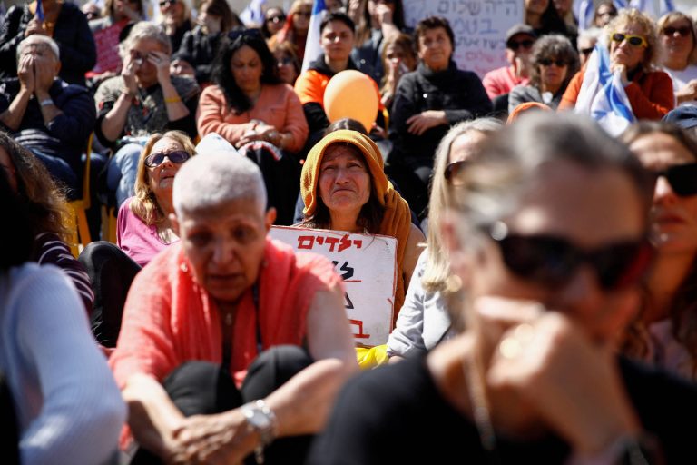 A imagem mostra um grupo de pessoas sentadas em um protesto. Em primeiro plano, há uma mulher com cabelo curto e grisalho, vestindo uma blusa vermelha e calças escuras. Ao fundo, uma mulher com um chapéu laranja e uma placa que contém texto em hebraico. O ambiente é de concentração, com várias pessoas ao redor, algumas com expressões sérias e outras olhando para frente.