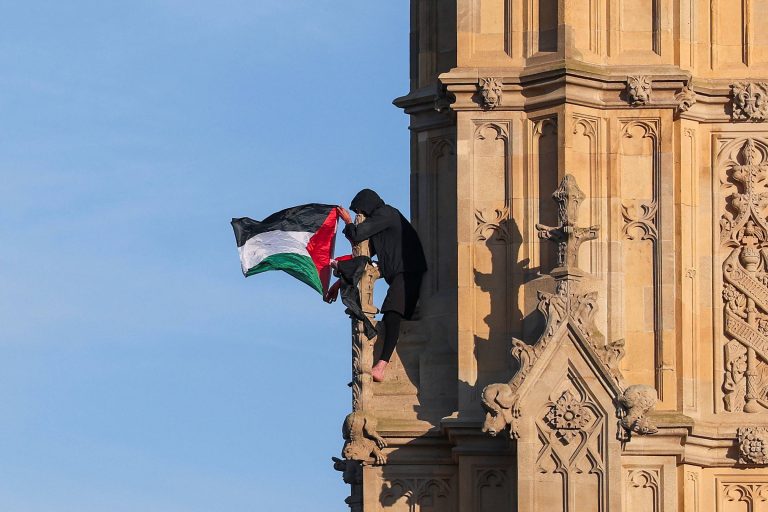 Uma pessoa está escalando uma torre de um edifício, segurando uma bandeira da Palestina, que possui as cores preta, branca, verde e vermelha. A pessoa está vestida de preto e parece estar posicionada em uma parte alta da estrutura, com um céu claro ao fundo.