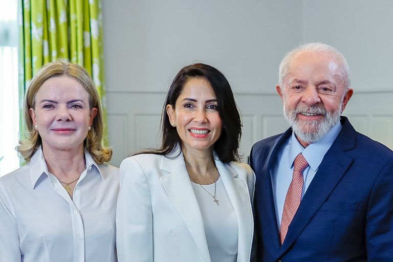 A imagem mostra três pessoas posando juntas em um ambiente interno. À esquerda, Gleisi, uma mulher com cabelo loiro e liso, vestindo uma camisa branca. No centro, Luisa Gonzalez, uma mulher com cabelo escuro e liso, usando um blazer branco e sorrindo. À direita, Lula, um homem idoso com cabelo grisalho e barba, vestido com um terno azul e uma gravata vermelha. Ao fundo, há cortinas verdes com padrões.