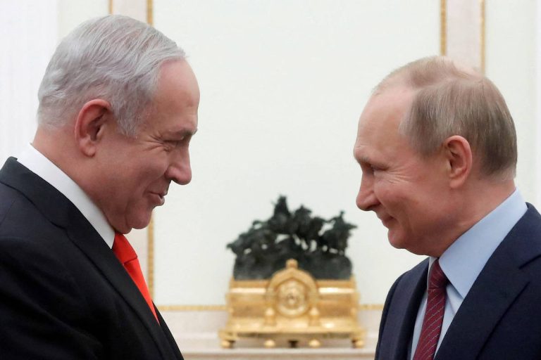 A imagem mostra dois homens em um encontro, sorrindo um para o outro. O homem à esquerda tem cabelo grisalho e usa um terno escuro com uma gravata vermelha. O homem à direita tem cabelo claro e usa um terno escuro com uma gravata clara. Ao fundo, há uma decoração simples com um objeto dourado e uma planta.