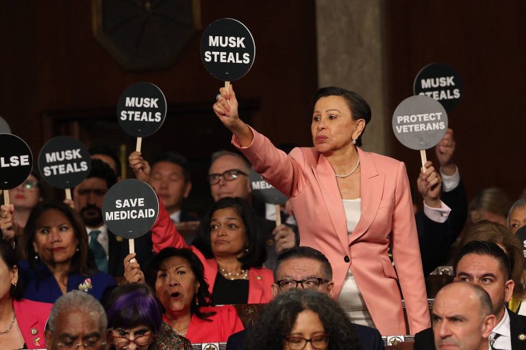 A imagem mostra um grupo de pessoas em uma audiência pública, segurando placas com mensagens de protesto. Uma mulher em destaque, vestindo um blazer rosa, levanta uma placa que diz