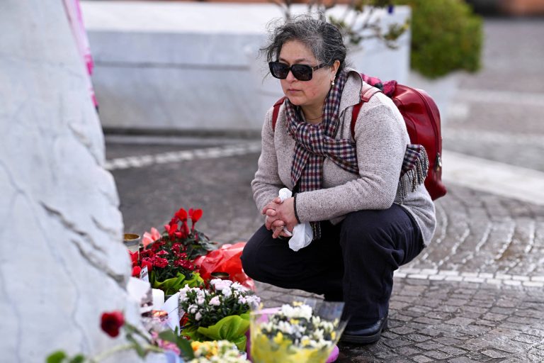 Uma mulher de cabelos grisalhos, usando óculos escuros e uma mochila vermelha, está agachada em frente a um memorial. Ela observa um local onde há flores e velas, demonstrando um momento de reflexão ou luto.