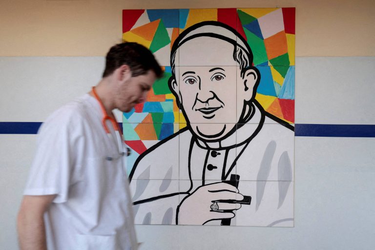 Um profissional de saúde, vestido com um jaleco branco e um estetoscópio no pescoço, caminha em frente a um mural colorido que retrata um homem com características que lembram um papa. O mural é composto por formas geométricas em várias cores, com o rosto do papa em destaque, mostrando um sorriso e segurando um crucifixo.