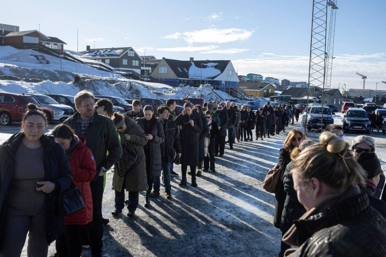 A imagem mostra uma longa fila de pessoas em um ambiente ao ar livre, com neve no chão e edifícios ao fundo. As pessoas estão vestidas com roupas de inverno, algumas segurando objetos. O céu está claro e ensolarado, e há sombras projetadas no chão devido à luz do sol. No fundo, é possível ver carros estacionados e uma torre de comunicação.
