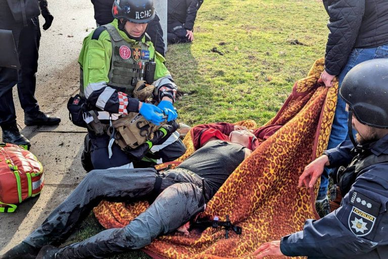 A imagem mostra uma cena de emergência onde vários socorristas estão prestando assistência a uma pessoa caída no chão. Um dos socorristas, vestido com um colete verde e capacete, está aplicando cuidados à vítima, que está coberta com um cobertor de pele de animal. Outros socorristas e policiais estão ao redor, observando e ajudando na situação.