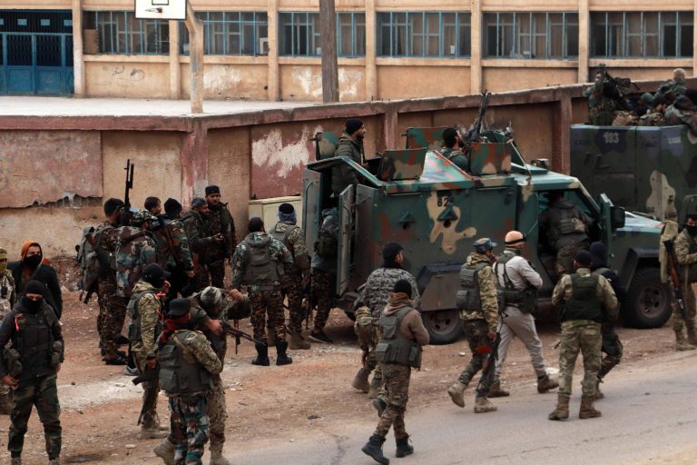 A imagem mostra um grupo de soldados armados em uma área urbana. Alguns soldados estão próximos a um veículo militar, enquanto outros estão em movimento. Ao fundo, há um prédio com janelas e uma quadra de esportes visível. O ambiente parece ser de um conflito, com a presença de equipamentos militares.