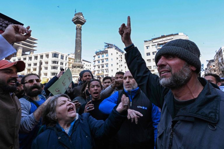 A imagem mostra um grupo de pessoas em um protesto em uma praça. No centro, um homem de barba e gorro levanta a mão, enquanto uma mulher à sua esquerda grita. Ao fundo, há um monumento e vários edifícios. O ambiente parece agitado, com várias pessoas expressando suas opiniões.