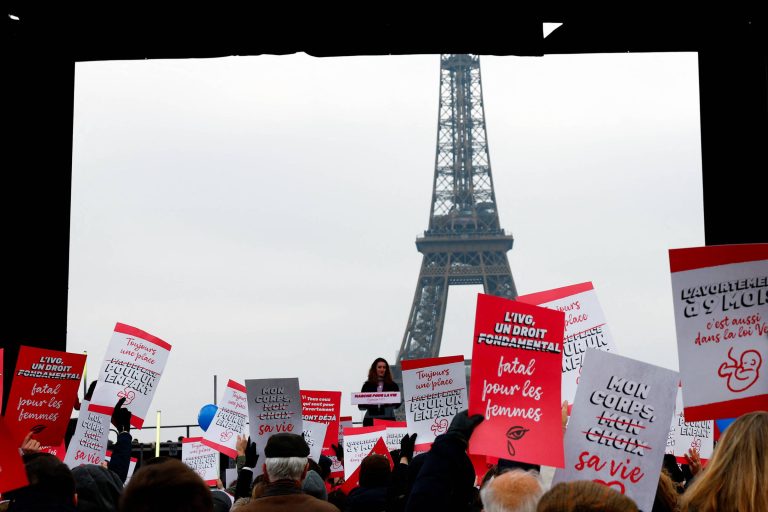 A imagem mostra uma manifestação em frente à Torre Eiffel, em Paris. Há uma multidão segurando cartazes vermelhos e brancos com textos em francês. No centro, uma mulher fala ao microfone, enquanto a Torre Eiffel se ergue ao fundo sob um céu nublado.
