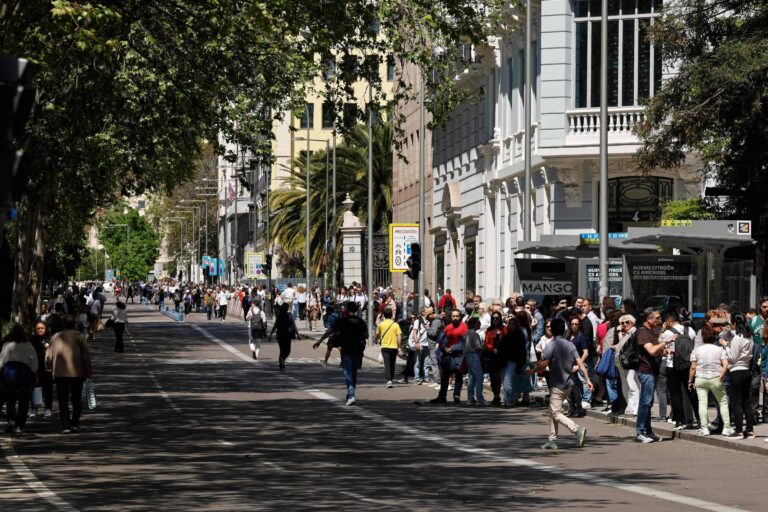A imagem mostra uma rua movimentada com muitas pessoas caminhando. À esquerda, há um grupo de pessoas em pé, enquanto à direita, uma fila de pessoas espera em um ponto de ônibus. O ambiente é urbano, com árvores e edifícios ao fundo, e a luz do sol ilumina a cena.