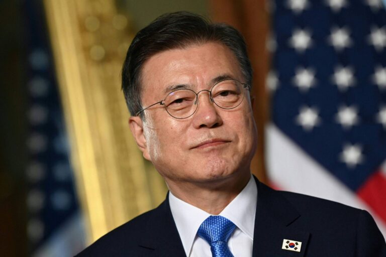 Um homem com cabelo escuro e óculos, vestindo um terno escuro e uma gravata azul. Ele está em um ambiente formal, com bandeiras dos Estados Unidos e da Coreia do Sul ao fundo. O homem tem uma expressão séria e está olhando diretamente para a câmera.