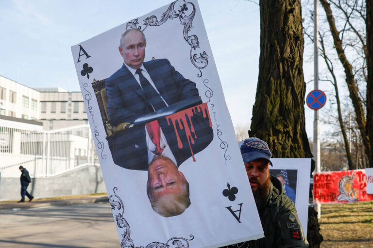 Um homem segura um cartaz que representa uma carta de baralho, com a figura de Vladimir Putin na parte superior e a de Donald Trump na parte inferior. A imagem de Putin está em uma posição normal, enquanto a de Trump está de cabeça para baixo. Há um efeito de sangue escorrendo da imagem de Putin. Ao fundo, é possível ver um edifício e outras pessoas participando do protesto.