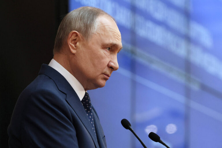 Vladimir Putin está de terno escuro em um palco, olhando para baixo, com um fundo desfocado que parece ser uma tela de apresentação. Ele tem cabelo curto e está usando uma gravata. O ambiente parece ser um evento formal, possivelmente uma conferência ou discurso.