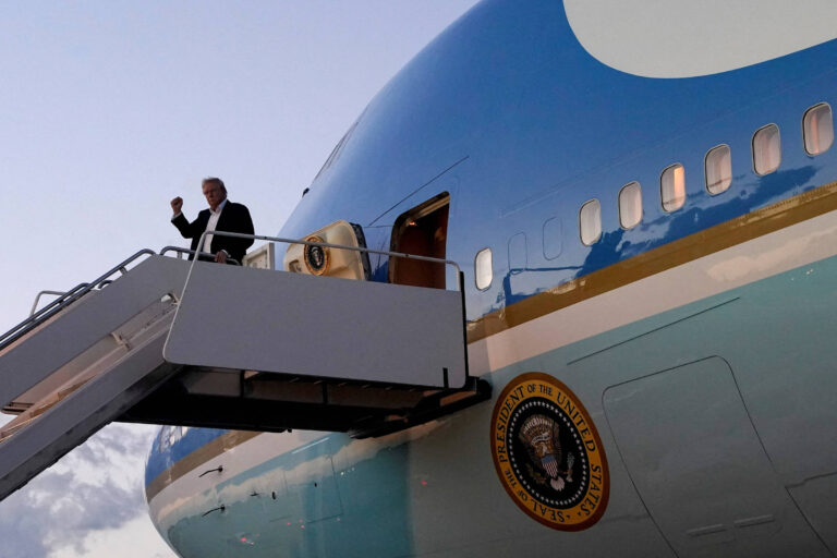 Um homem está saindo de um avião, que é identificado como Air Force One, através de uma escada. Ele está acenando com a mão. O avião é de cor azul e branca, com o emblema da presidência dos Estados Unidos visível na parte inferior da fuselagem. O céu está claro, indicando que é um momento do dia.