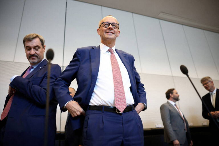 A imagem mostra um grupo de homens em um ambiente de conferência. Um homem em destaque, vestido com um terno azul e gravata vermelha, está sorrindo e ajustando seu paletó. Outros homens estão ao fundo, alguns com expressões neutras, todos vestidos formalmente. O ambiente parece ser um auditório ou sala de reuniões, com microfones visíveis na frente.