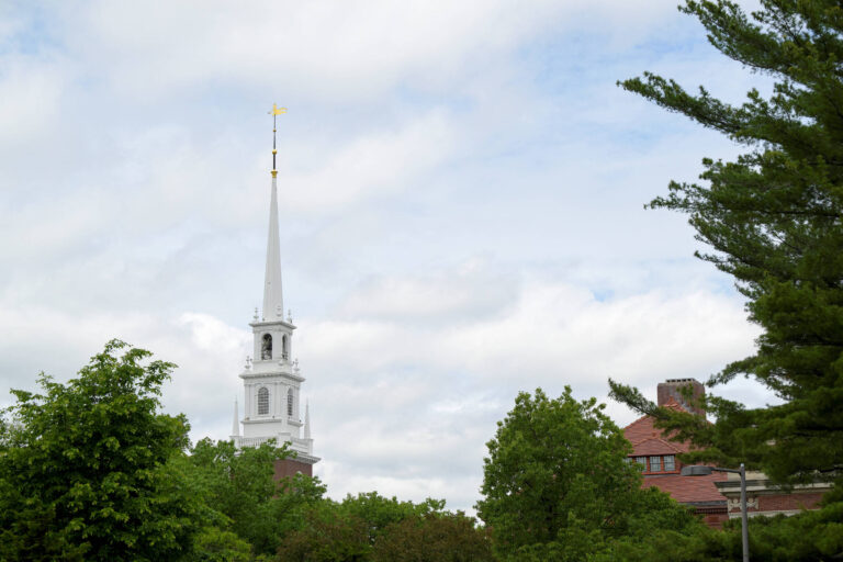 A imagem mostra uma igreja com uma torre alta e esguia, que se destaca contra um céu nublado. A torre possui uma cruz dourada no topo. Ao redor da igreja, há árvores verdes e um edifício de tijolos visível à direita.