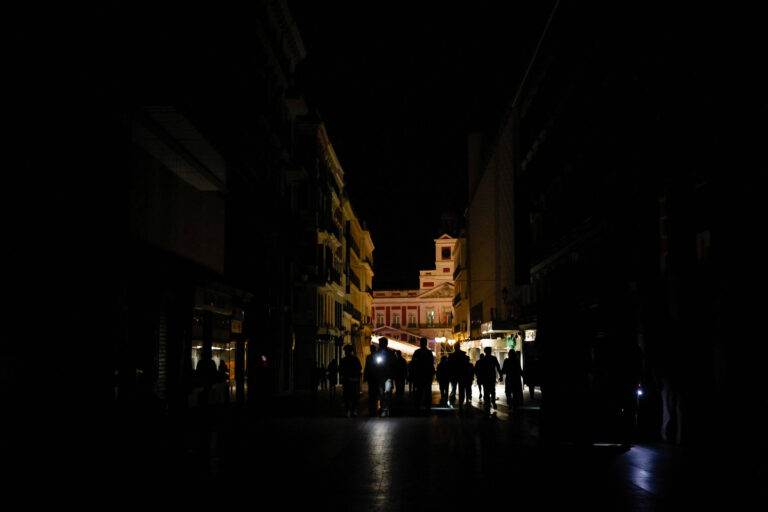 A imagem mostra uma rua escura à noite, com algumas pessoas caminhando. Ao fundo, há uma construção iluminada, possivelmente um edifício ou uma praça, que se destaca na escuridão. As sombras das pessoas são visíveis, e algumas delas estão segurando lanternas ou celulares, emitindo luz. As fachadas dos prédios ao redor são pouco iluminadas, criando um contraste com a luz do fundo.