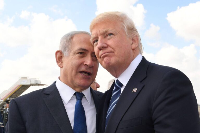 A imagem mostra dois homens em um ambiente ao ar livre, aparentemente conversando de forma próxima. Um deles está vestindo um terno escuro com uma gravata clara, enquanto o outro usa um terno escuro com uma gravata azul. Ao fundo, é possível ver uma aeronave e um céu com nuvens.