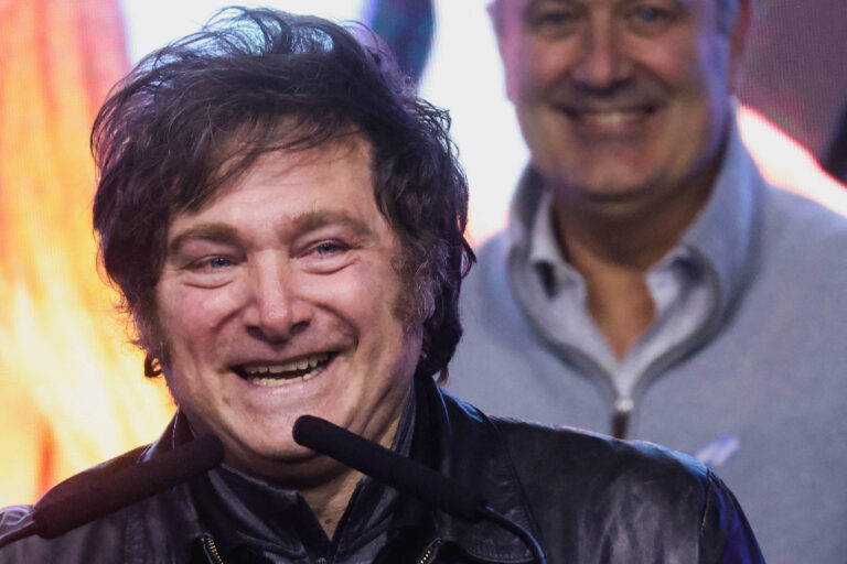 A imagem mostra um homem sorridente com cabelo bagunçado, usando uma jaqueta preta. Ele está em um palco, com microfones à sua frente, e parece estar se dirigindo a uma plateia. Ao fundo, há um homem sorrindo, que parece estar prestando atenção ao palestrante. O ambiente é iluminado e colorido, sugerindo um evento animado.