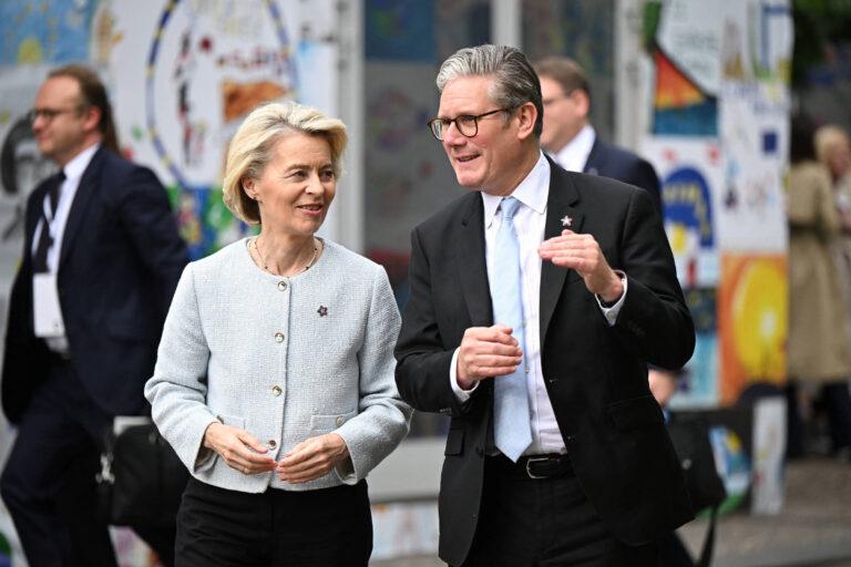 Duas pessoas estão caminhando lado a lado em um ambiente externo. A mulher à esquerda tem cabelo loiro e usa um blazer claro, enquanto o homem à direita usa um terno escuro e óculos. Ambos parecem estar conversando de forma amigável. Ao fundo, há uma parede decorada com várias pinturas e ilustrações coloridas, e outras pessoas podem ser vistas ao longe.